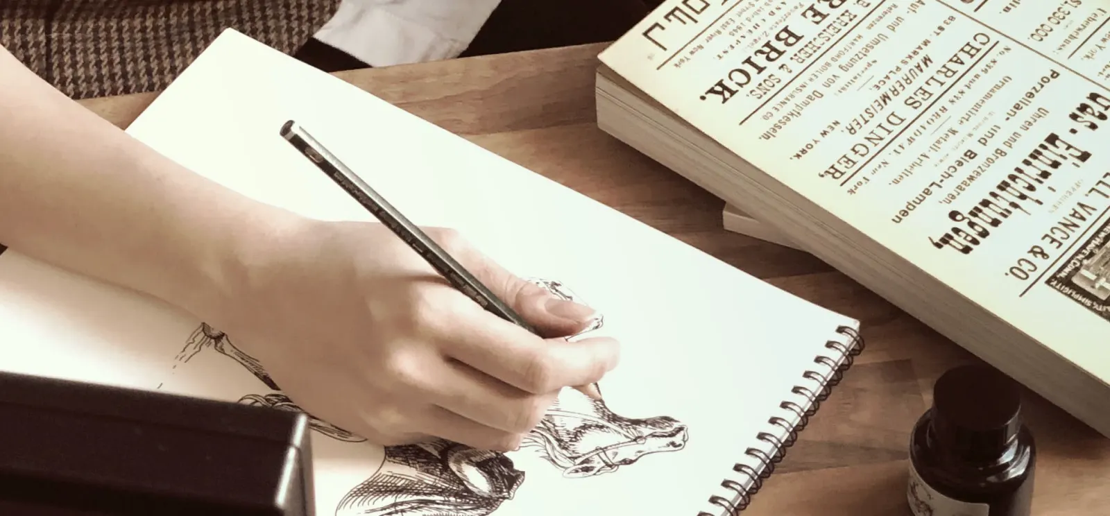 A hand sketching a creature in a sketchbook beside an inkwell.