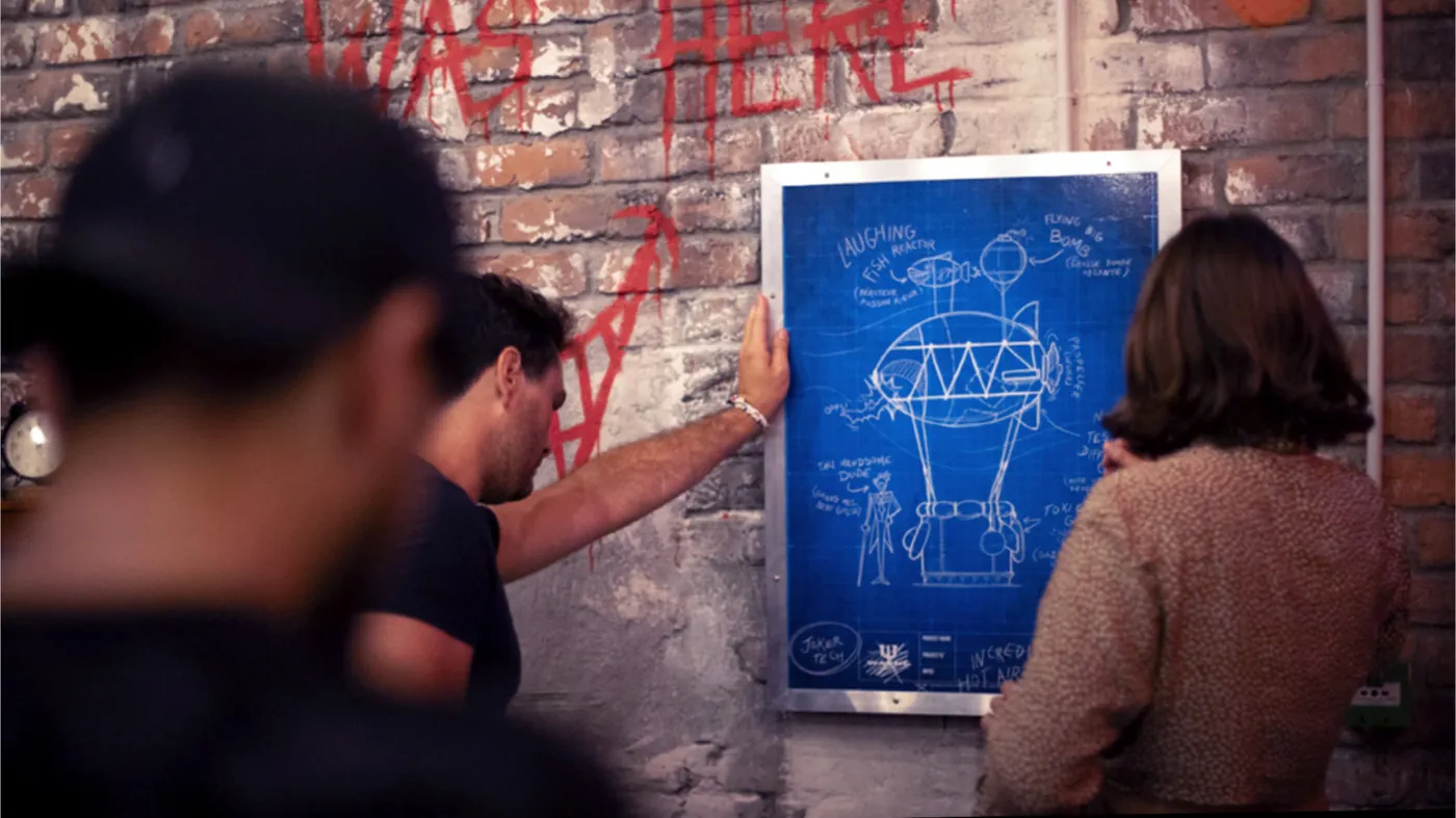 Two people studying a blueprint of a Joker contraption pinned to a brick wall.