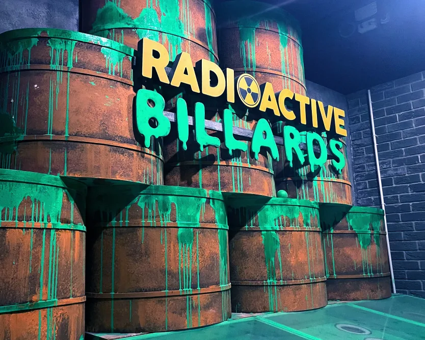 Radioactive Billiards neon sign glowing green above stacked toxic barrels.