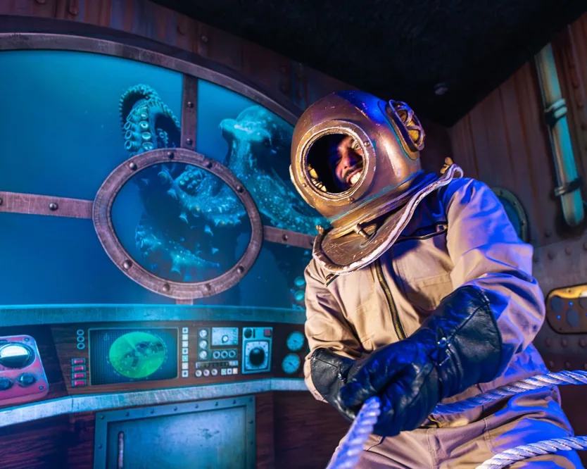 A diver in a vintage brass-helmet suit at the controls of an immersive submarine set.